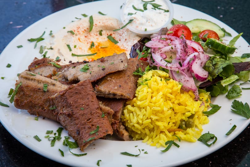 A gyro platter with sliced meat, yellow rice, salad, hummus, and tzatziki sauce.