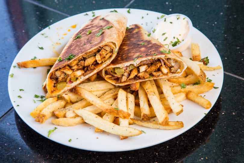 Two chicken shawarma wraps, cut in half, served with french fries and a side of white sauce.