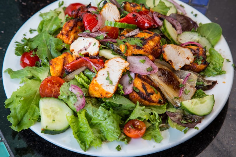 Grilled chicken and vegetable salad with lettuce, tomatoes, cucumbers, and red onions on a white plate.