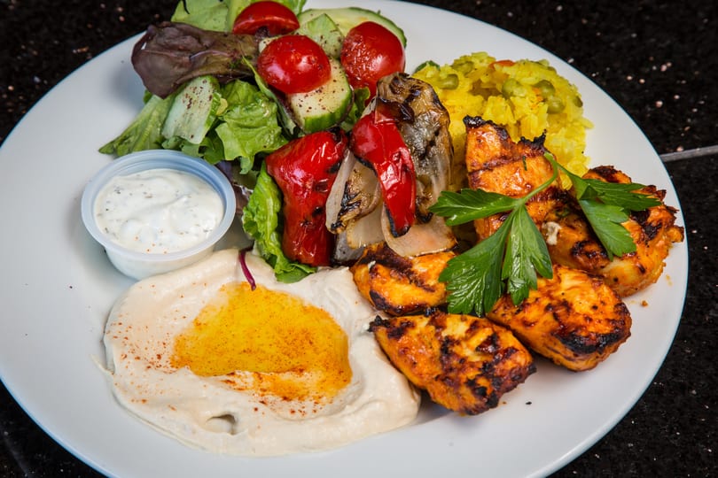 A plate of grilled chicken, hummus, rice, salad, and roasted vegetables with a side of sauce.